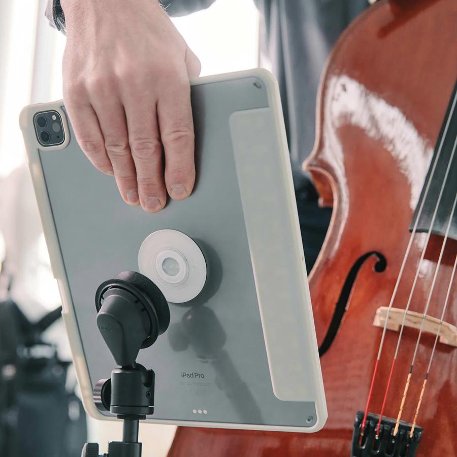 Hand hält das Tablet um es mit die runden VACUUM-Verbindung am Notenständer zu befestigen, daneben ist ein Cello zu sehen.